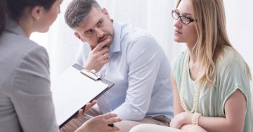 A man and woman look tense as they sit in front of a woman with a clipboard.