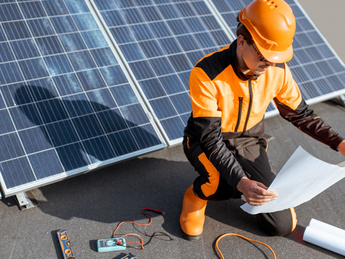 An electrician kneeling next to solar panels on a roof and looking at blueprints.