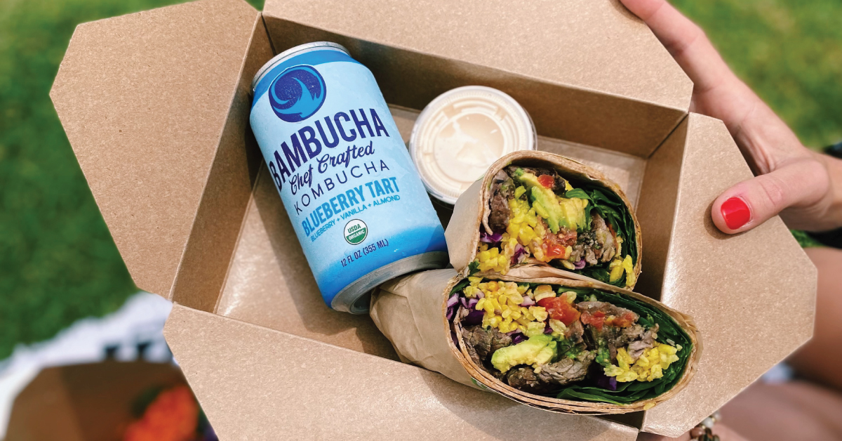 hands holding box with wrap and kombucha at a picnic
