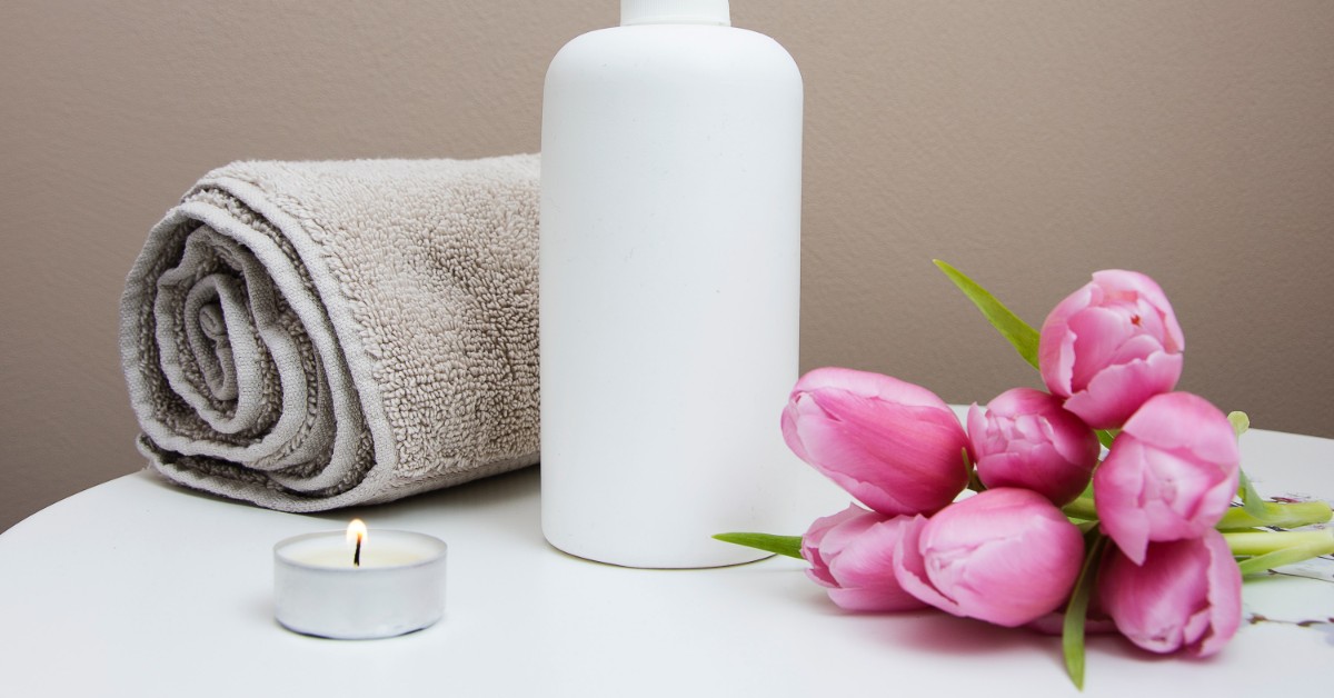 Rolled up towel, lit candle, pink flowers, and a cleanser.